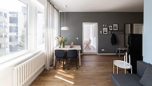 A modern living room with a grey sofa, a dining area with a table and chairs, and a large window with curtains. - Greystay Apartments Steglitz (Berlin)