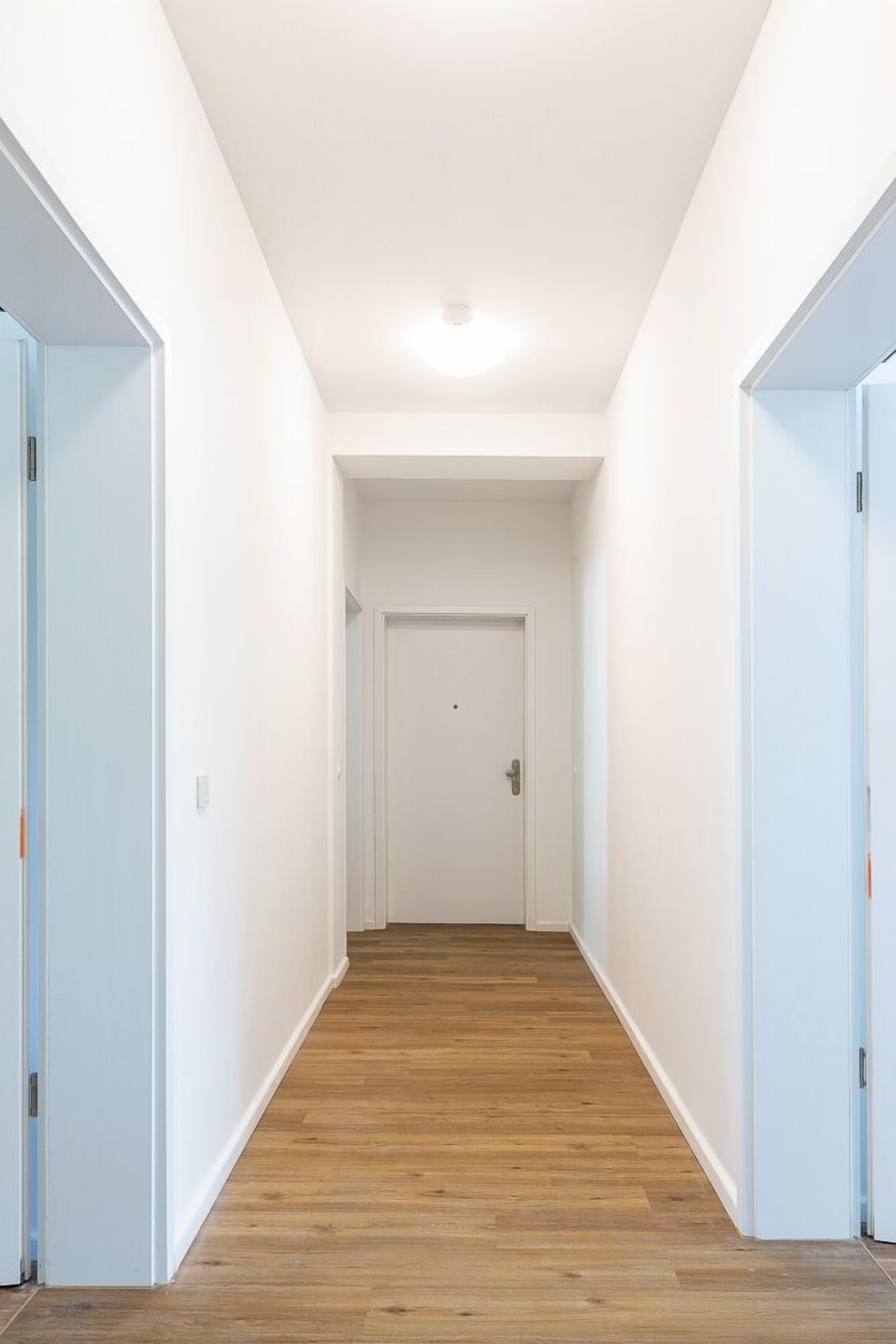 A narrow hallway with wooden flooring and white walls, leading to a closed door at the end.