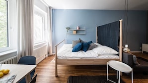 A modern bedroom with a wooden bed, a desk with a chair, and a window with curtains. - Greystay Apartments Steglitz (Berlin)