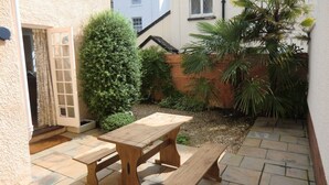 Eine Terrasse mit Holztisch und Bänken, umgeben von einem Garten mit verschiedenen Pflanzen und einer Backsteinmauer.