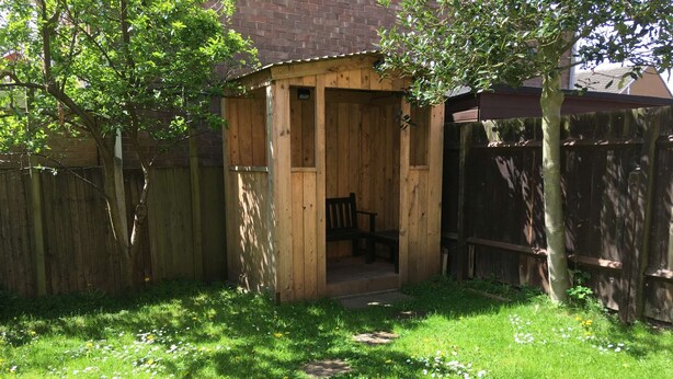 A wooden shed with a bench inside, surrounded by a wooden fence and a grassy yard with scattered flowers.