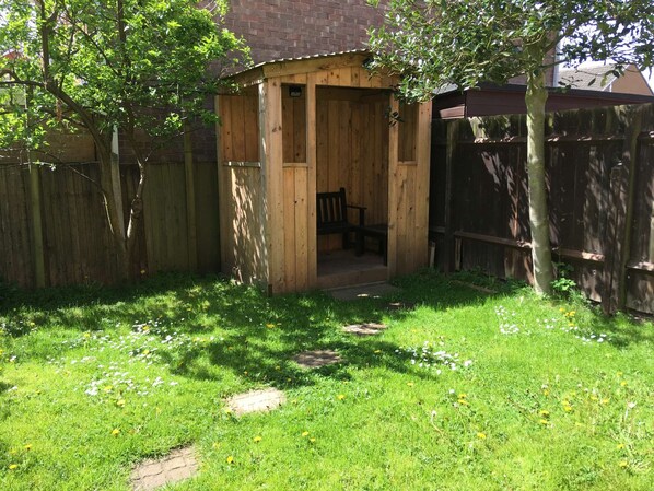 A wooden shed with a bench inside, surrounded by a wooden fence and a grassy yard with scattered flowers.