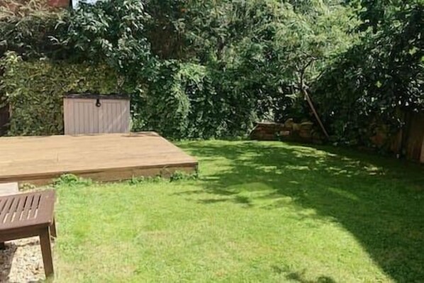 Een houten terras met een bankje, omgeven door een verzorgd gazon en weelderige begroeiing.