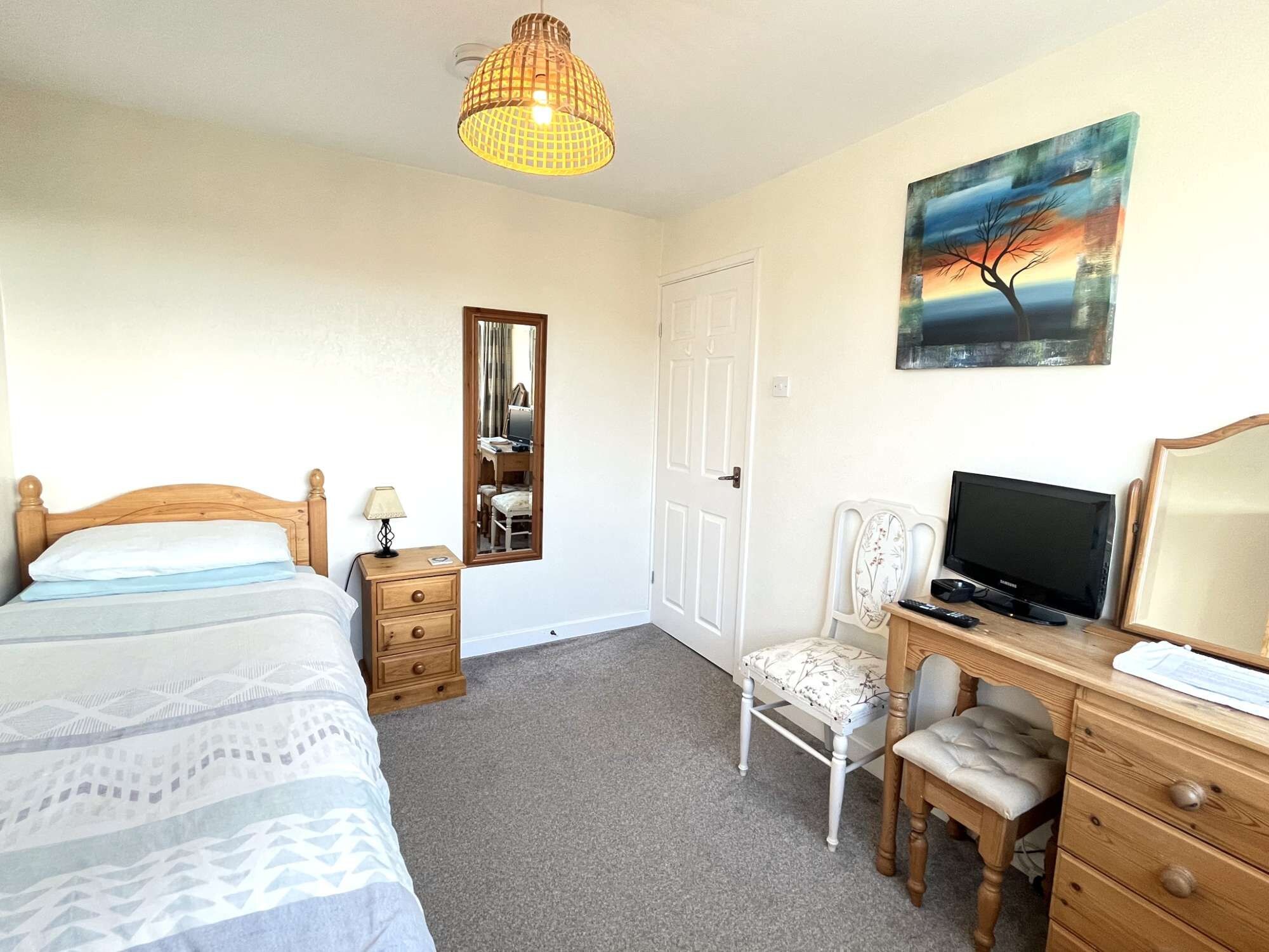 A bedroom with a bed, bedside table, lamp, chair, desk with a TV, and a painting on the wall.