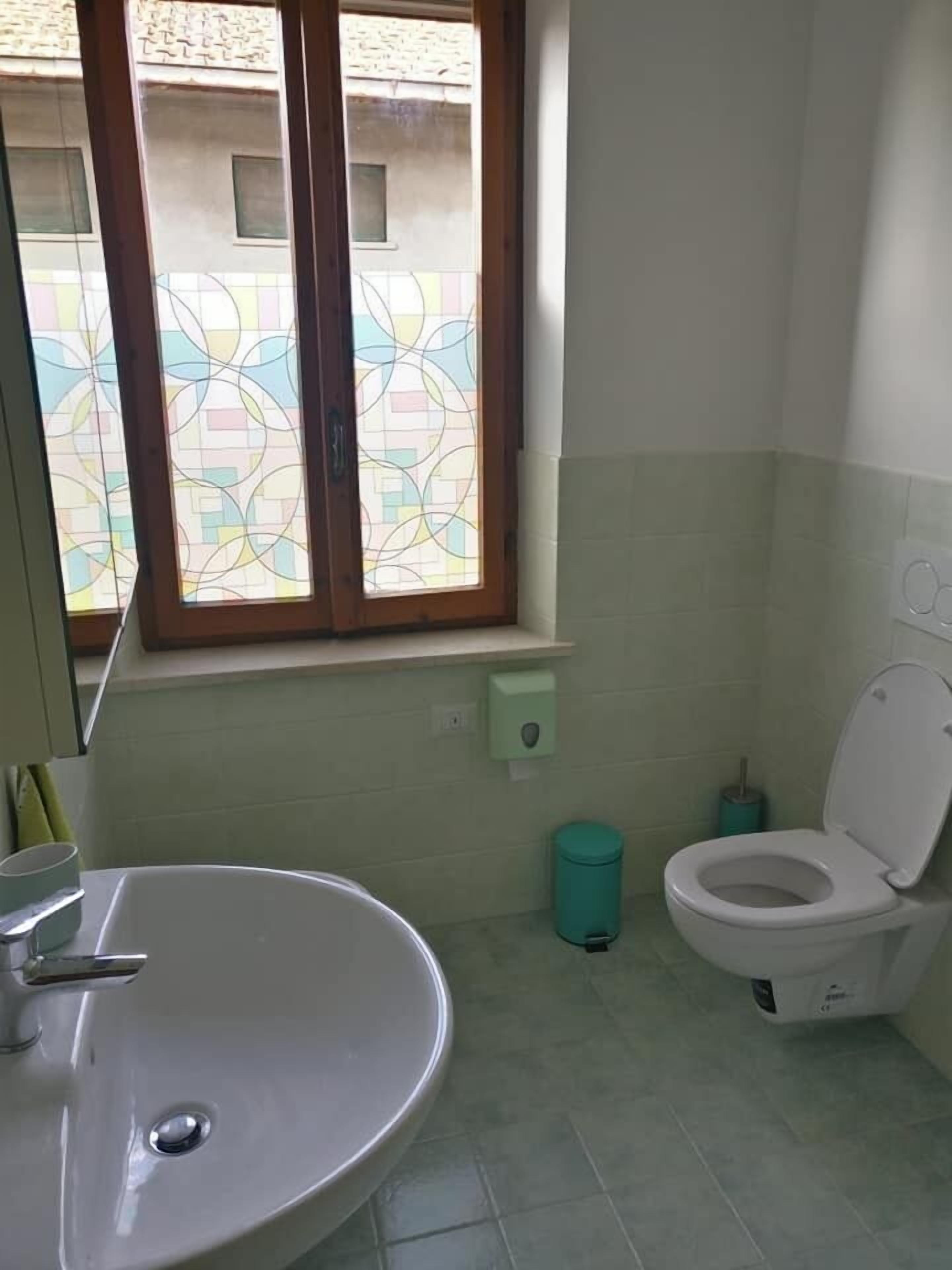A bathroom with a white sink, a toilet, a window with a geometric stained glass design, and a green trash bin.