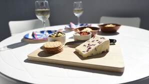 Una mesa redonda con mantel blanco, tabla de madera para cortar, un bloque de queso, un tazón de aceitunas, un tazón de nueces, un plato de galletas y dos copas de vino blanco.