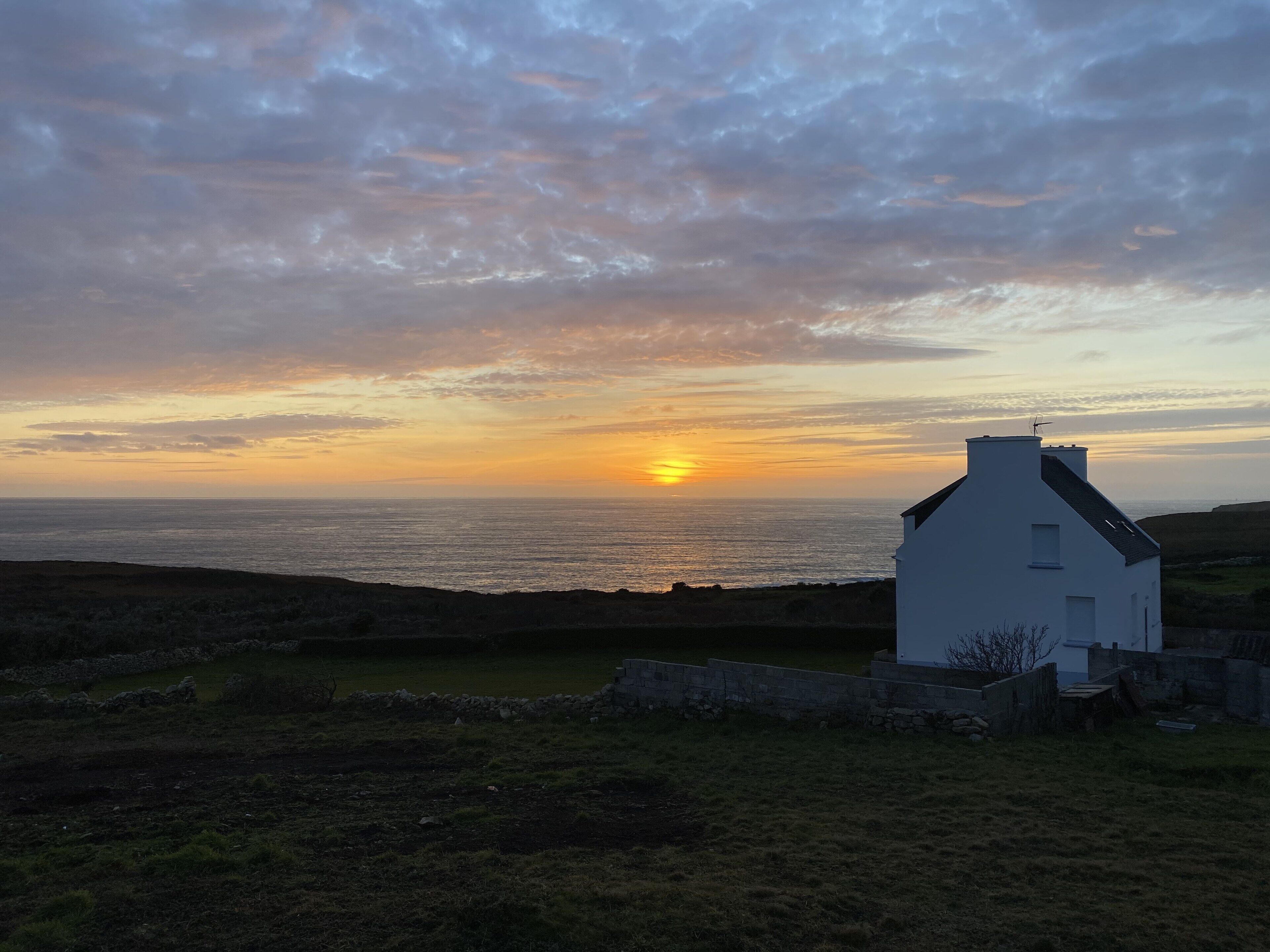 A white house with a stone wall in front, overlooking a body of water during sunset.
