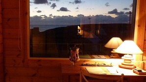 Habitació de cabana de fusta amb una finestra gran que fa vedes a un paisatge muntanyós. Hi ha un escriptori de fusta amb una làmpada, una cadira i alguns papers.