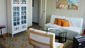 Un salon avec un canapé blanc, des coussins orange, une table basse en verre, un meuble blanc avec des portes en verre et un tableau coloré accroché au mur.