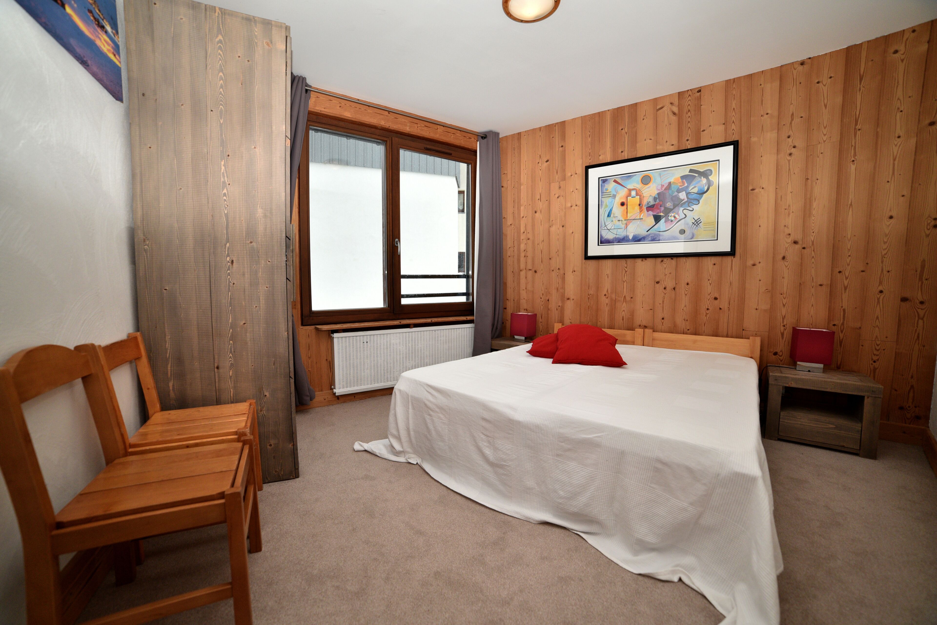 A bedroom with a wooden paneled wall, a large bed with a white bedspread and a red pillow, a wooden chair, a nightstand with a red lamp, and a window with a view of the outside.