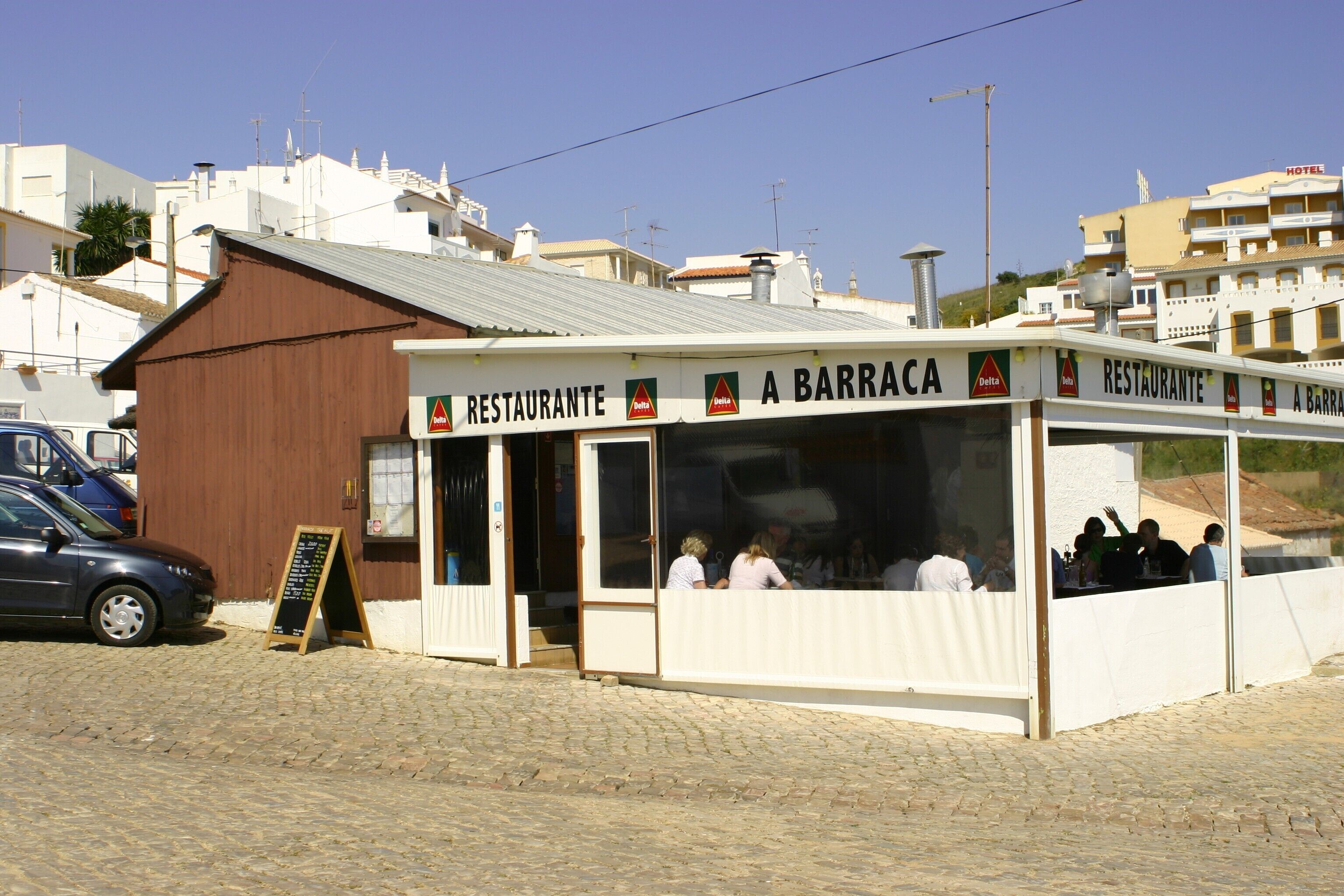 Un restaurant situé sur la côte, avec une terrasse, un menu affiché de manière visible et une voiture garée à proximité.
