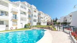 Una piscina rodeada de edificios blancos con balcones, un camino pavimentado y un cielo azul despejado.