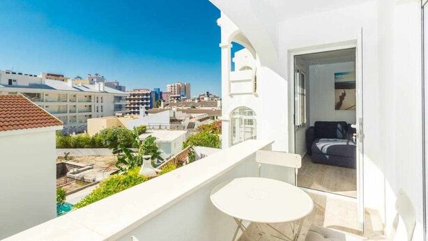 Un balcón con una mesa y sillas blancas, con vistas a un paisaje urbano con edificios y un cielo azul despejado.