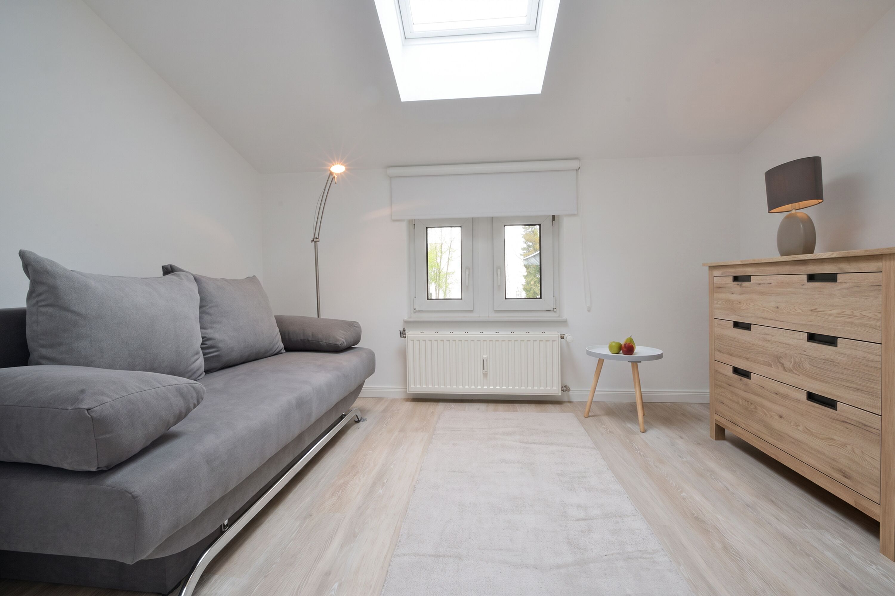 Una sala de estar moderna con un sofá gris, un aparador de madera, una mesita con fruta y un tragaluz arriba.