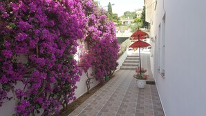 Un estrecho callejón con una pared cubierta de flores de buganvilla moradas, un paraguas rojo y una maceta con flores rosadas.