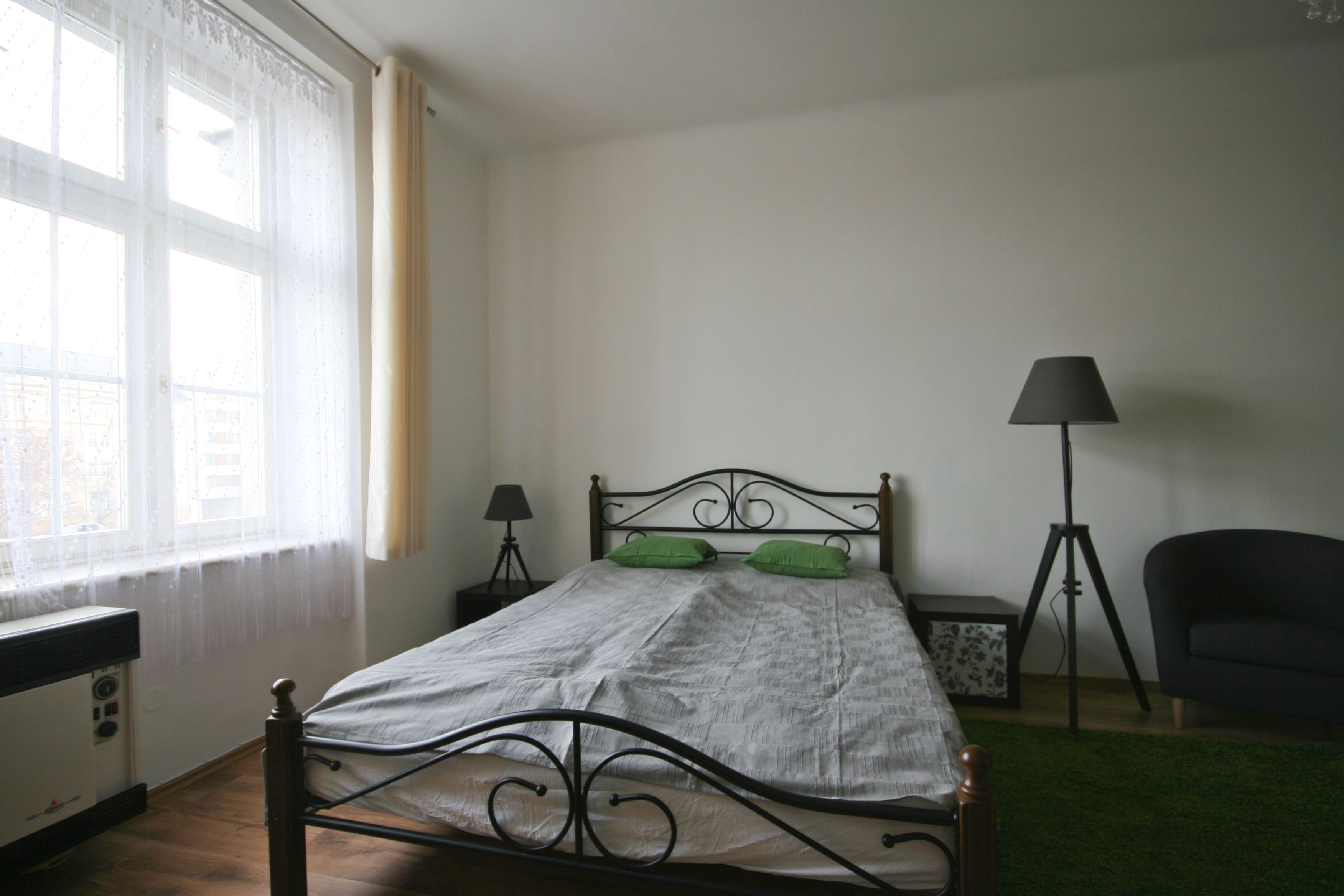 A bedroom with a bed, a window with sheer curtains, a bedside table with a lamp, and a chair.