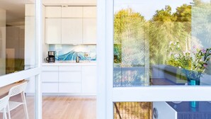 Een moderne keuken met witte kastjes en een koffiezetapparaat, een zithoek met een witte stoel, en een balkon met uitzicht op bomen en bloemen.