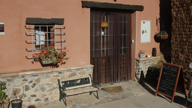 A building with a stone facade, a wooden bench, a metal window with a flower basket, and a chalkboard sign. - El Tío Carrascón - Hostel (Cerveruela)