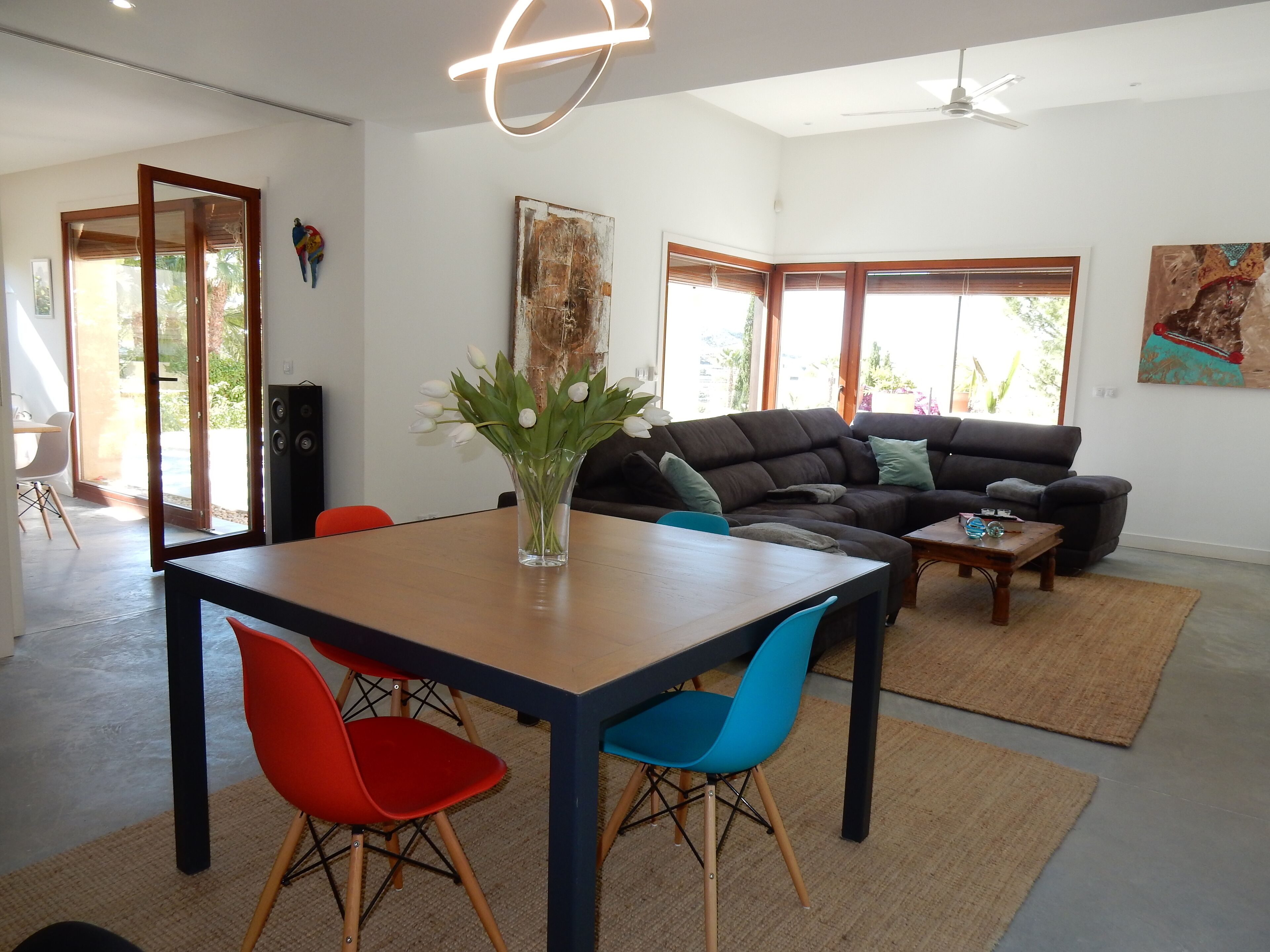 A modern living room with a dark-colored sofa, a wooden coffee table, and a dining table with red and blue chairs. There is a vase with white flowers on the dining table.