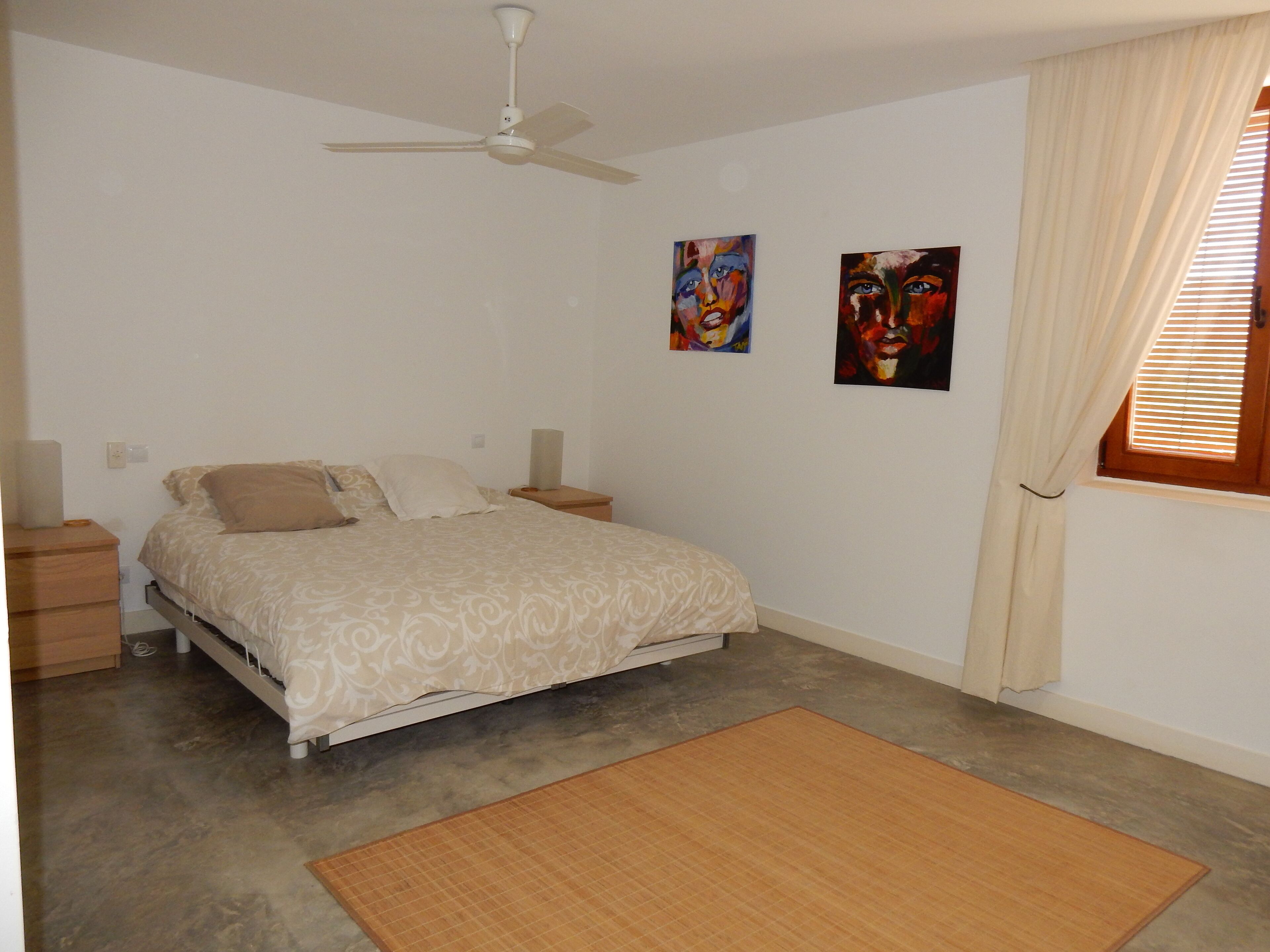 A bedroom with a bed, nightstands, a ceiling fan, two paintings, a window with blinds, and a mat on the floor.