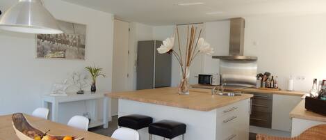 A modern kitchen with a central island, white chairs, a wooden dining table, and a hanging pendant light.