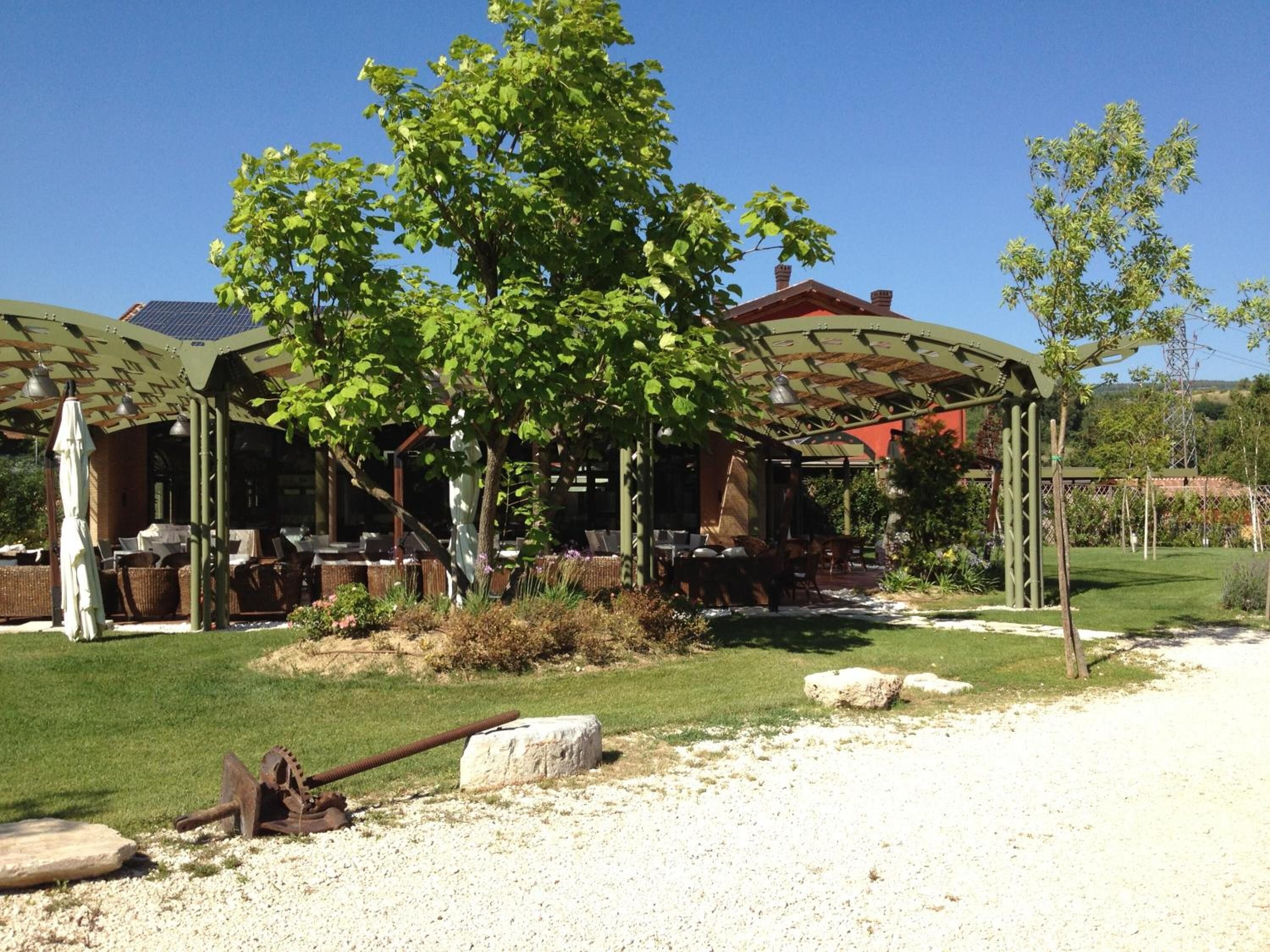 A covered outdoor dining area with tables and chairs, surrounded by greenery and a gravel path.