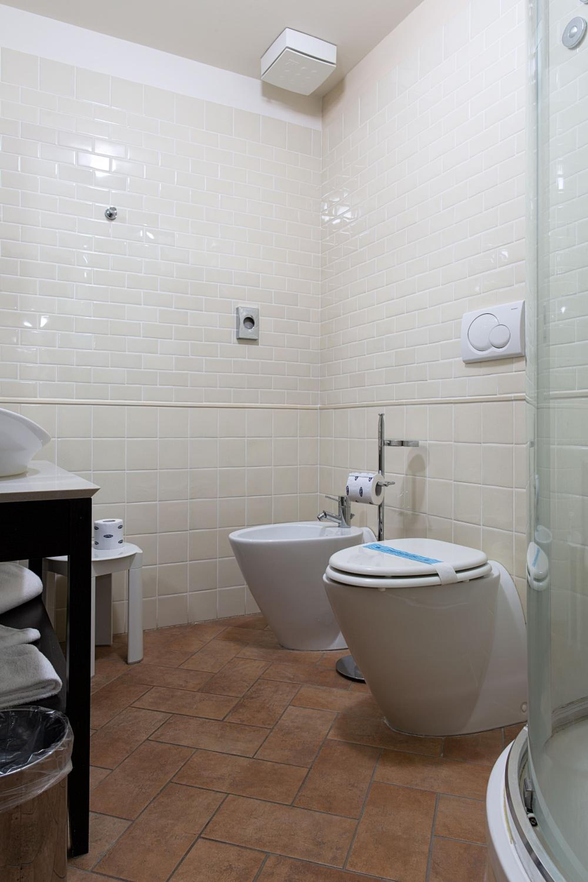 Un baño con paredes de azulejos blancos, un inodoro, un bidé, un lavamanos y un cubo de basura.