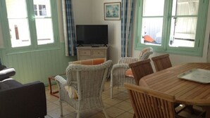 A dining area with wicker chairs and a wooden table, a television on a dresser, and a window with curtains.