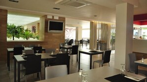 Un restaurante moderno con techo alto, iluminación empotrada y una pantalla de televisión grande. Las mesas están dispuestas con sillas, y se ve un jardín a través de un ventanal.