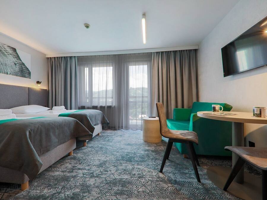 Habitación de hotel con dos camas, un escritorio con silla, un televisor y un ventanal con cortinas.