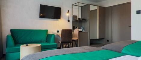 A hotel room with a green sofa, a desk, a chair, a bed with green and grey bedding, and a TV mounted on the wall.