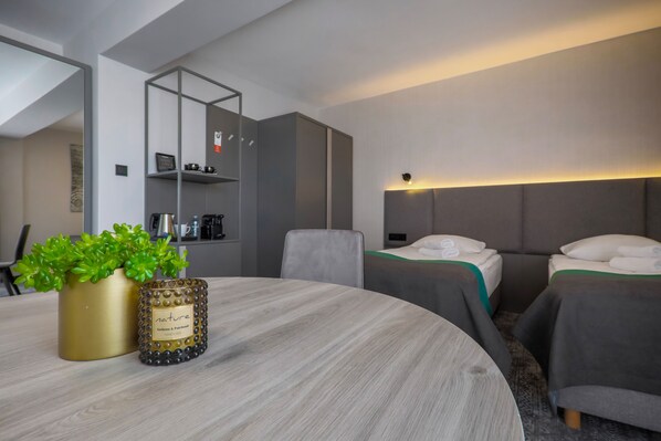 Una habitación de hotel moderna con una mesa redonda, dos camas, un sofá, una pequeña cocina y una planta en maceta.