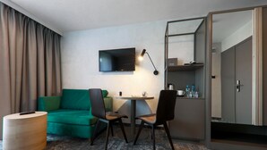 Quarto de hotel com um sofá verde, duas cadeiras, uma mesinha, TV na parede e um minibar.