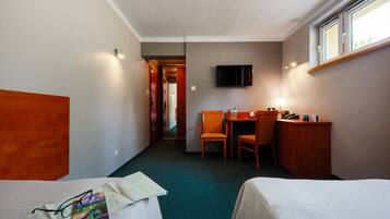 A hotel room with two single beds, a desk with a chair, a TV mounted on the wall, and a window providing natural light.