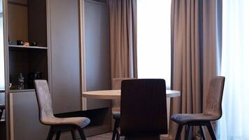 A hotel room with a dining area featuring a round table and four chairs, a minibar, and a large window with curtains.