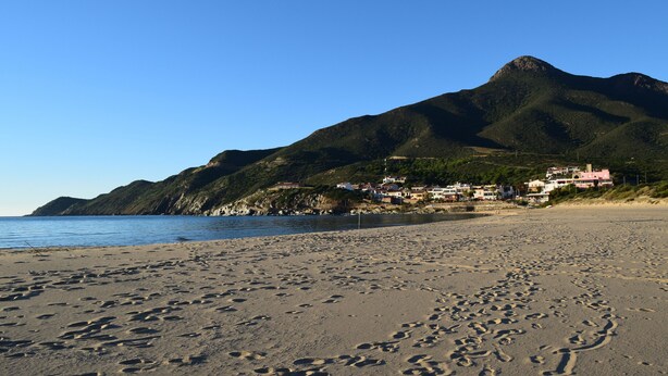 A beach with footprints leading to the water, a mountain in the background, and buildings on the hillside. - Stylishly designed apartment with magnificent sea views in a stunning natural environment (Portixeddu)
