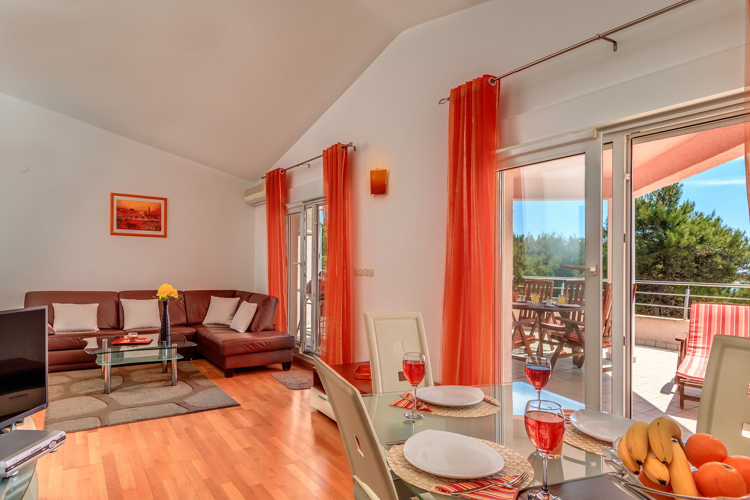 Ein modernes Wohnzimmer mit braunem Sofa, Glastisch und einem Essbereich mit Glastisch, roten Vorhängen sowie einem Balkon mit Gartenmöbeln.