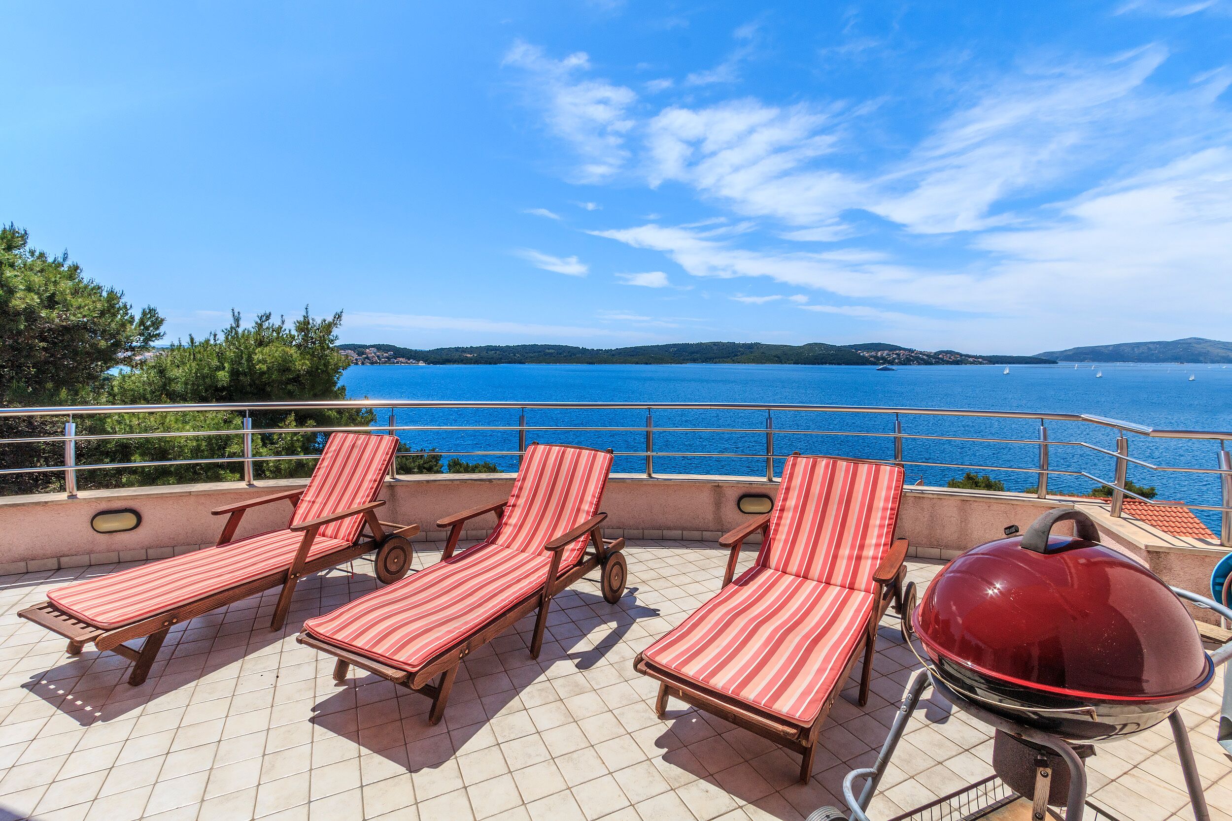 A terrace with four striped lounge chairs and a barbecue grill, overlooking a body of water and hills.