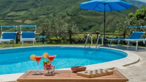 A poolside setting with a view of green hills, a blue umbrella, and a table with drinks and a chessboard.