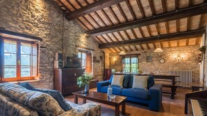 A rustic living room with stone walls, wooden beams, a blue sofa, a floral-patterned sofa, a coffee table, and a piano.