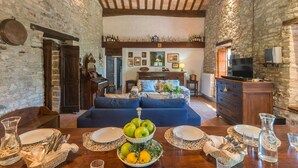 A rustic living room with stone walls, a wooden ceiling, a sofa, and a dining table set with plates, glasses, and a fruit bowl.