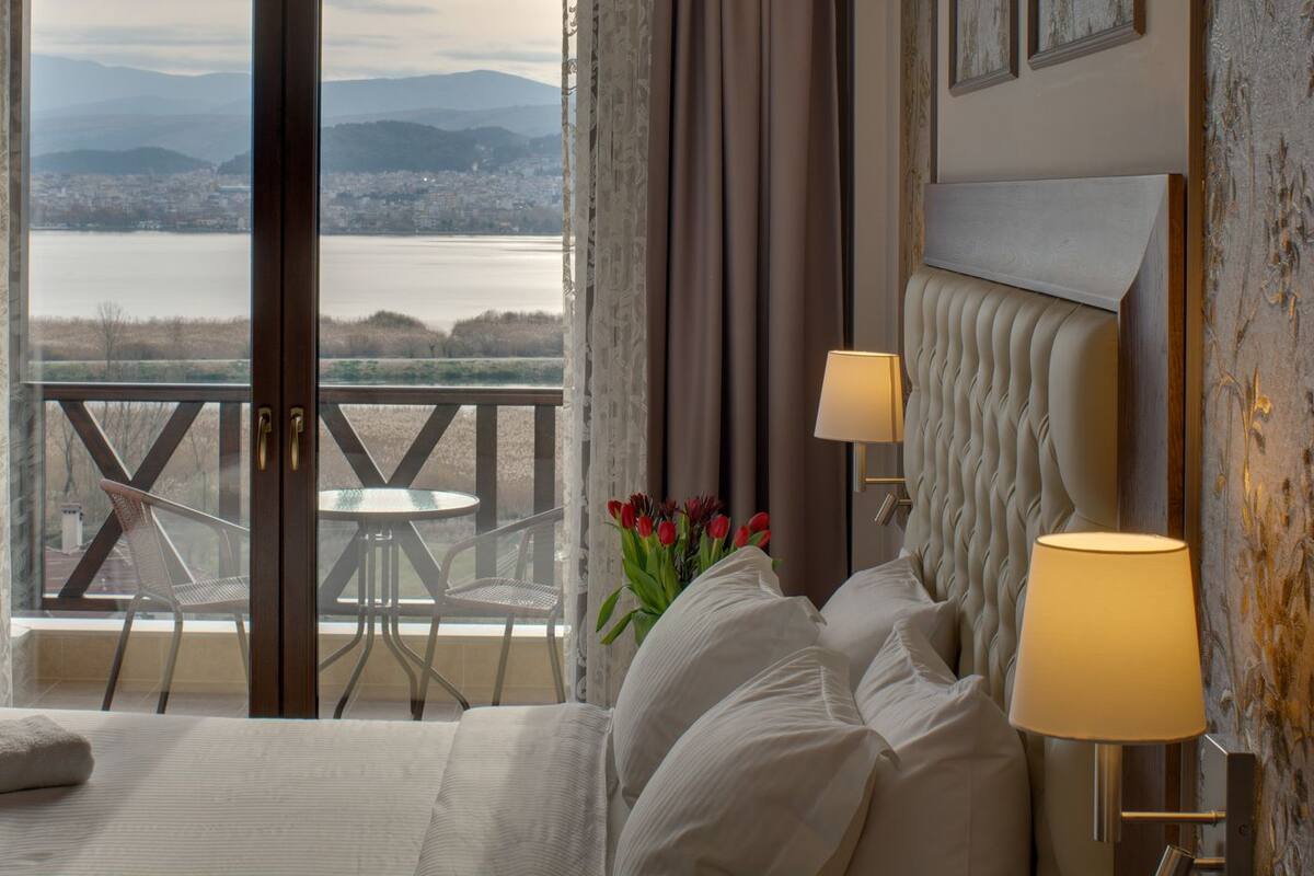 Ein Hotelzimmer mit Bett, zwei Nachttischlampen, einem Fenster mit Blick auf Stadt und Berge, einem Balkon mit Tisch und Stühlen sowie einer Vase mit roten Tulpen.