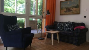 Un salon avec un fauteuil bleu foncé, un canapé à carreaux, une petite table en bois et un lampadaire. Il y a une grande fenêtre avec des rideaux orange et un tableau accroché au mur.