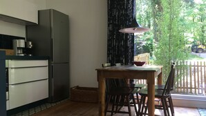 Une cuisine compacte équipée d’un réfrigérateur, d’une table à manger avec des chaises, et offrant une vue sur un jardin par une fenêtre.
