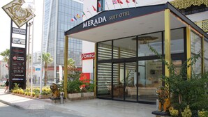Exterior del Hotel Merada Suite con fachada de vidrio, una estatua de león en primer plano y varias banderas sobre la entrada.