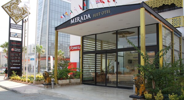 Exterior del Hotel Merada Suite con fachada de vidrio, una estatua de león en primer plano y varias banderas sobre la entrada.