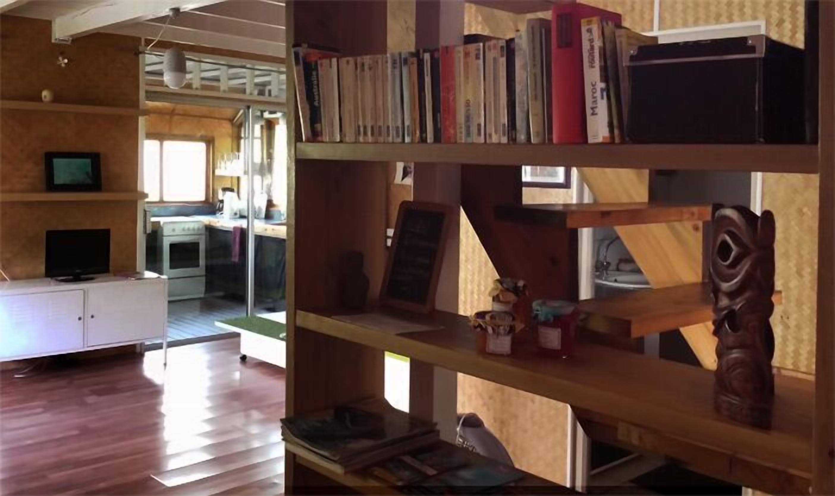 Um quarto com piso de madeira, um armário branco com televisão e uma estante cheia de livros e objetos decorativos.