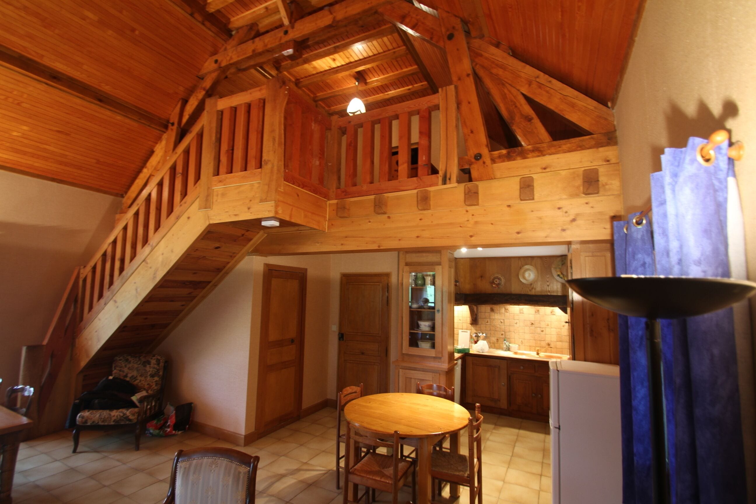 A spacious loft-style room with a high ceiling, wooden beams, a dining area with a wooden table and chairs, and a kitchen area with wooden cabinets and a sink.