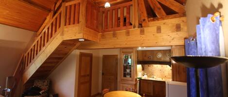 A spacious loft-style room with a high ceiling, wooden beams, a dining area with a wooden table and chairs, and a kitchen area with wooden cabinets and a sink.