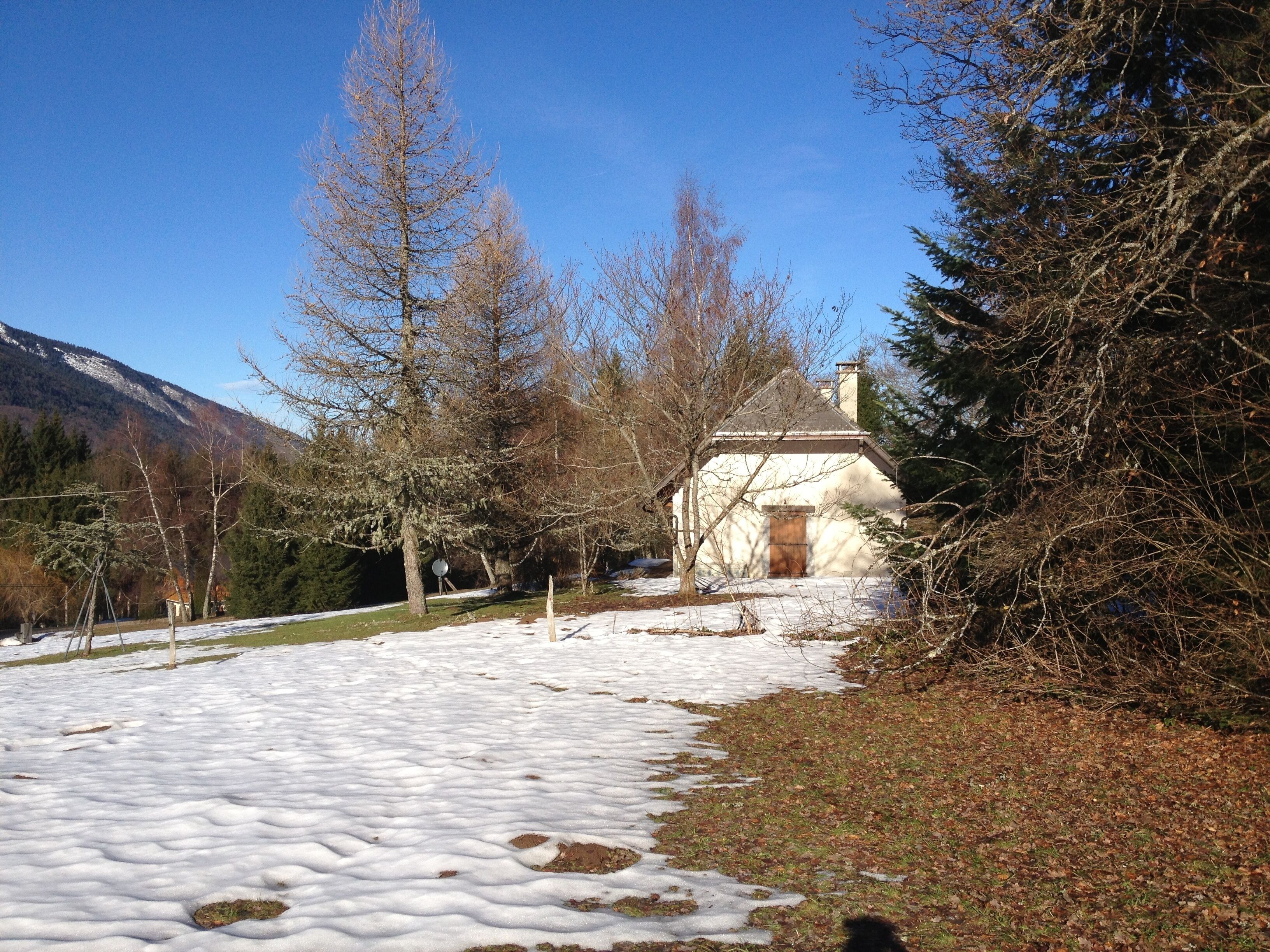 Sentiero innevato che conduce a una casa rustica con camino, circondata da alberi spogli e una montagna sullo sfondo.